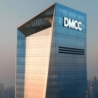 A tall, modern skyscraper with "DMCC" on top, set against a hazy city skyline at dusk.