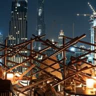 People dining on a rooftop terrace with modern skyscrapers and cranes illuminated against the night sky in the background.