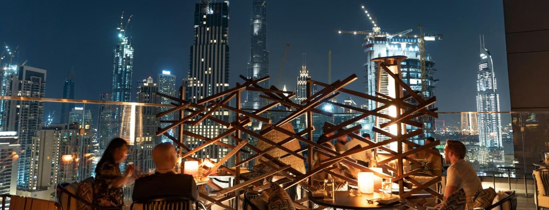 People dining on a rooftop terrace with modern skyscrapers and cranes illuminated against the night sky in the background.