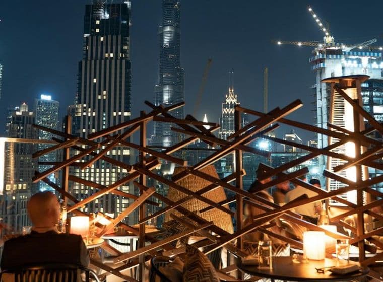 People dining on a rooftop terrace with modern skyscrapers and cranes illuminated against the night sky in the background.
