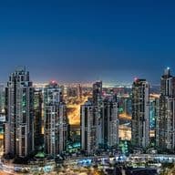 Panoramic view of a city skyline at night, featuring illuminated skyscrapers and the distinctive silhouette of a tall tower in the background.