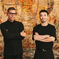 Zwei Männer stehen mit verschränkten Armen vor einem kunstvollen, farbenfrohen Wandteppich. Beide tragen dunkle Kleidung und Brillen und blicken selbstbewusst in die Kamera.