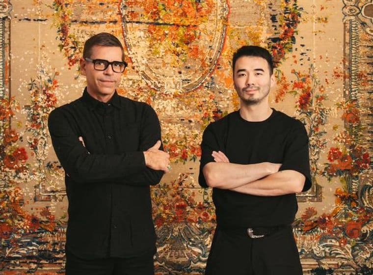 Zwei Männer stehen mit verschränkten Armen vor einem kunstvollen, farbenfrohen Wandteppich. Beide tragen dunkle Kleidung und Brillen und blicken selbstbewusst in die Kamera.