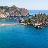 Panoramablick auf eine atemberaubende sizilianische Bucht mit kristallklarem, türkisfarbenem Wasser. In der Mitte der Bucht erhebt sich ein kleines felsiges Eiland, umgeben von festgemachten Booten. Die Küste ist von steilen, mit mediterraner Vegetation bewachsenen Felsklippen geprägt. Im Hintergrund erstreckt sich das Meer in tiefen Blautönen bis zum Horizont. Der weiß-graue Kieselstrand liegt am Fuße der Felsen und bildet einen eindrucksvollen natürlichen Kontrast.