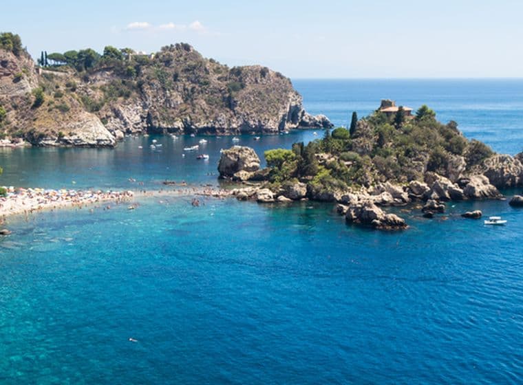 Panoramic view of a stunning Sicilian bay with crystal-clear turquoise waters. A small rocky islet rises at the center of the bay, surrounded by moored boats. The coastline is characterized by steep rocky cliffs covered in Mediterranean vegetation. In the background, the sea stretches to the horizon in deep blue hues. The white and gray pebble beach lies at the foot of the rocks, creating a striking natural contrast.