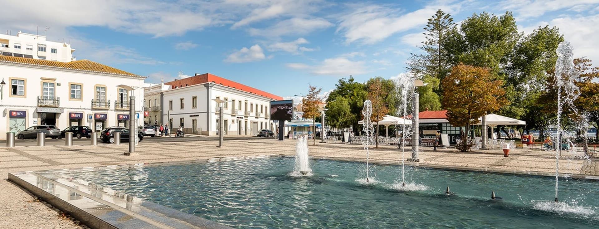 A public square with a fountain, surrounded by historic buildings, trees, and a clear blue sky in the background.