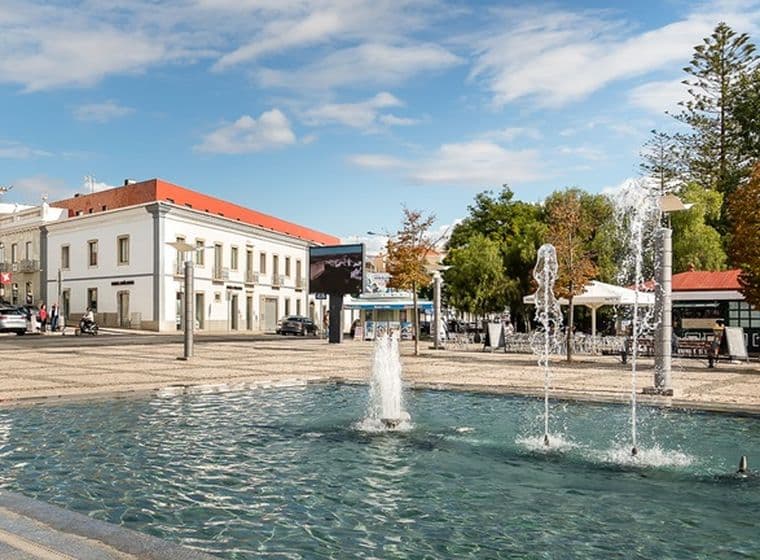 Ein öffentlicher Platz mit einem Brunnen, umgeben von historischen Gebäuden, Bäumen und einem klaren blauen Himmel im Hintergrund.