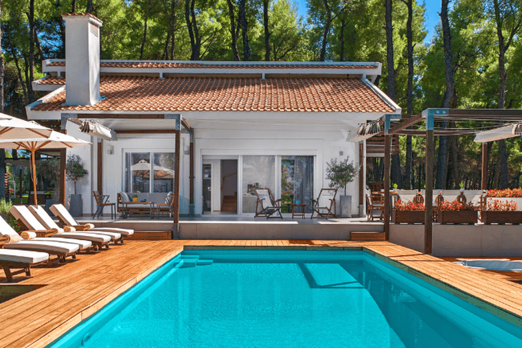 Luxury villa with a private swimming pool surrounded by pine trees. The property features a white exterior, terracotta roof tiles, a spacious wooden deck, sun loungers with umbrellas, and outdoor lounge areas, creating a tranquil resort-style setting.