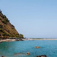 Küstenlandschaft mit einem felsigen Hügel, einer kurvenreichen Straße und einer Burg auf dem Gipfel, die ein ruhiges blaues Meer unter einem klaren Himmel überblickt.