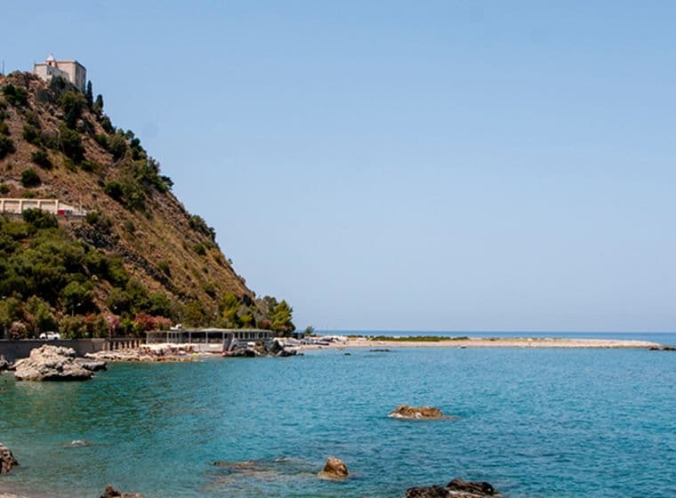Coastal landscape with a rocky hill, winding road, and a castle on top, overlooking a calm blue sea under a clear sky.
