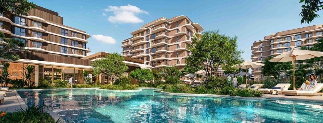 Luxurious apartment complex with modern buildings, lush greenery, and a large pool surrounded by lounge chairs and umbrellas under a clear blue sky.