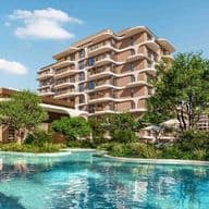 Luxurious apartment complex with modern buildings, lush greenery, and a large pool surrounded by lounge chairs and umbrellas under a clear blue sky.
