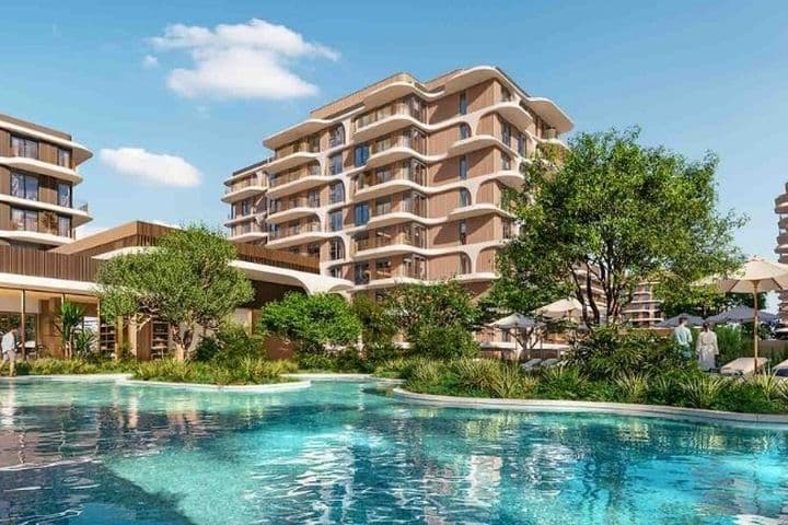 Luxurious apartment complex with modern buildings, lush greenery, and a large pool surrounded by lounge chairs and umbrellas under a clear blue sky.