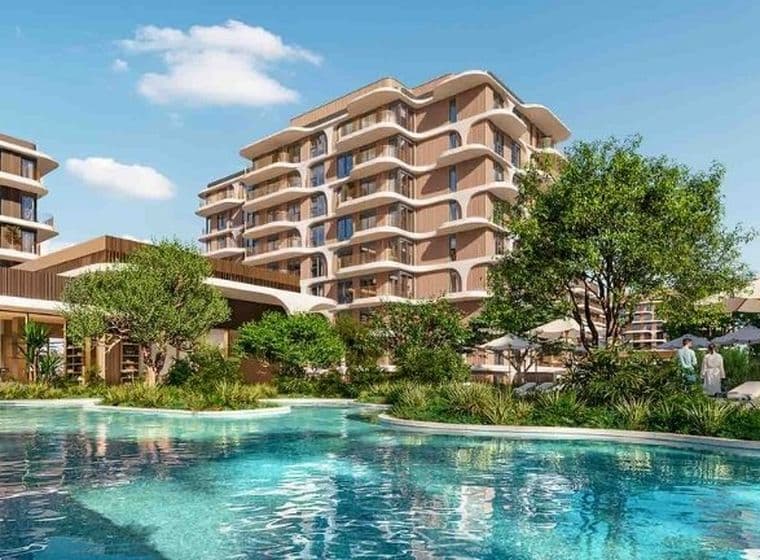 Luxurious apartment complex with modern buildings, lush greenery, and a large pool surrounded by lounge chairs and umbrellas under a clear blue sky.