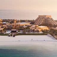 Aerial view of a luxury beachfront resort with pyramid-shaped buildings, surrounded by pools and palm trees, overlooking a calm sea at sunset.