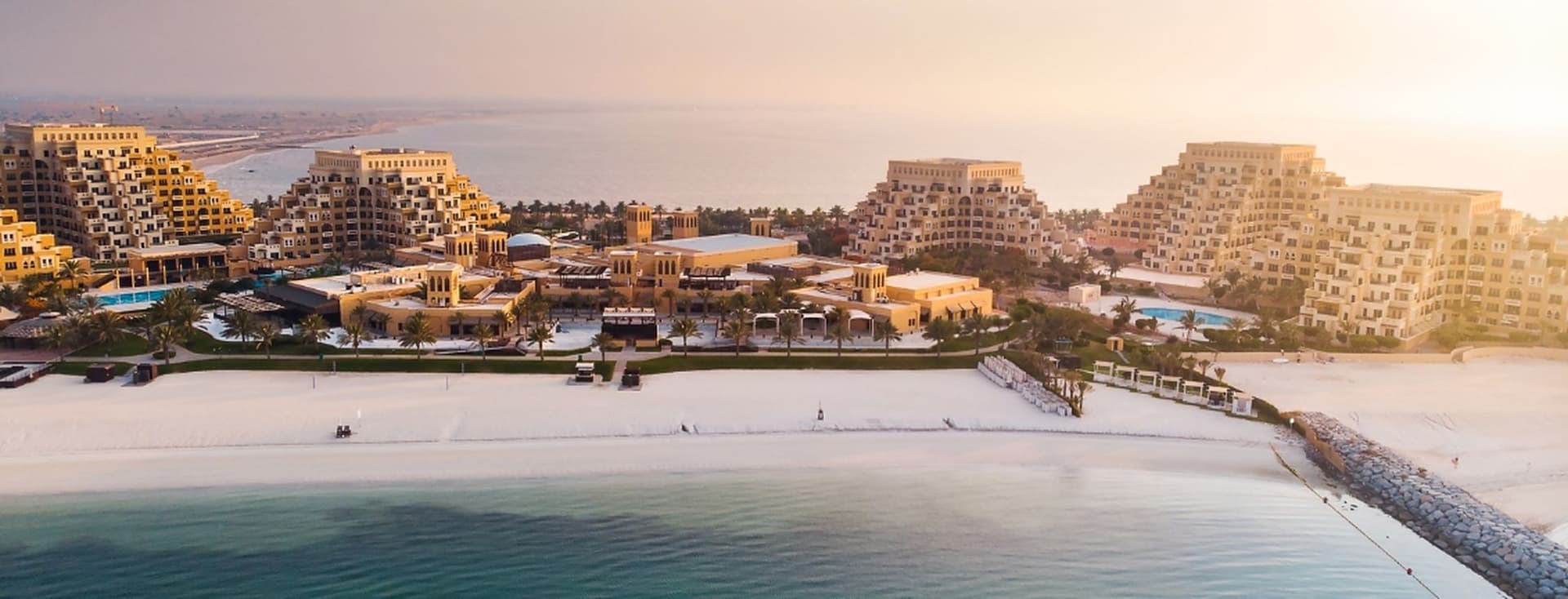 Aerial view of a luxury beachfront resort with pyramid-shaped buildings, surrounded by pools and palm trees, overlooking a calm sea at sunset.