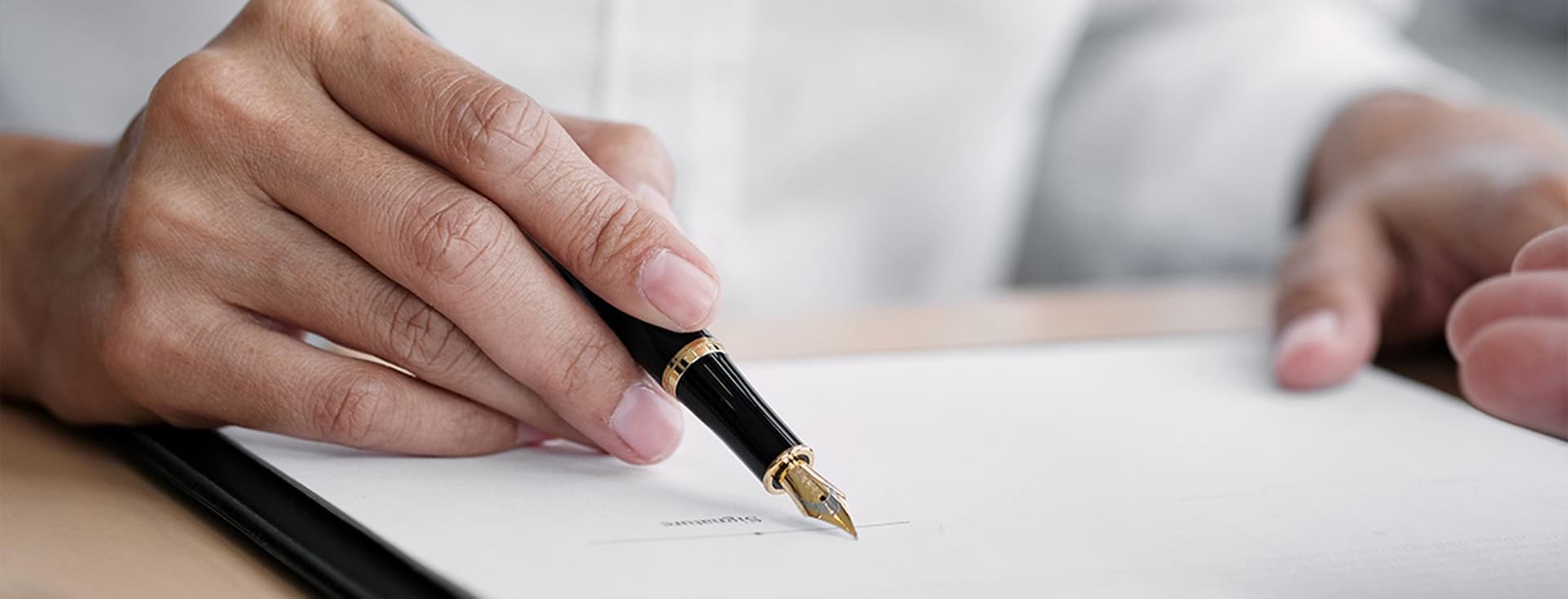 Hands holding a fountain pen poised over a document, ready to sign. The person is wearing a white shirt.