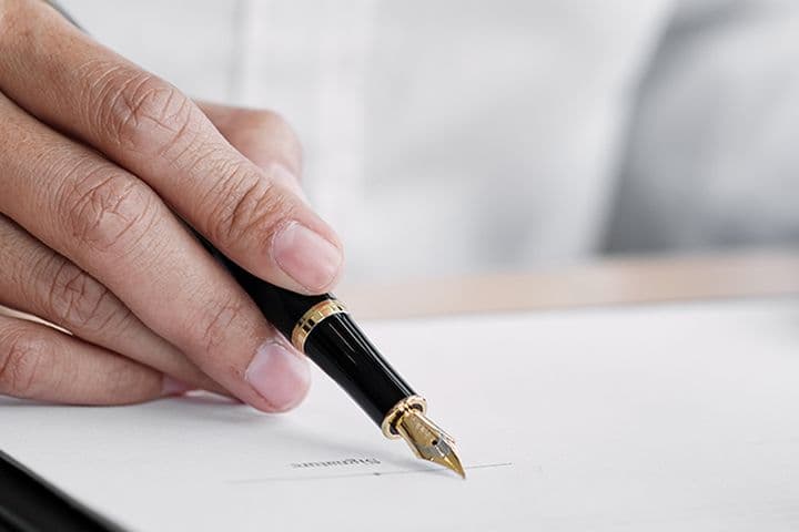 Hands holding a fountain pen poised over a document, ready to sign. The person is wearing a white shirt.