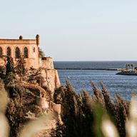 A historic castle on a cliff overlooks a calm sea, with a distant harbor and buildings under a clear blue sky.