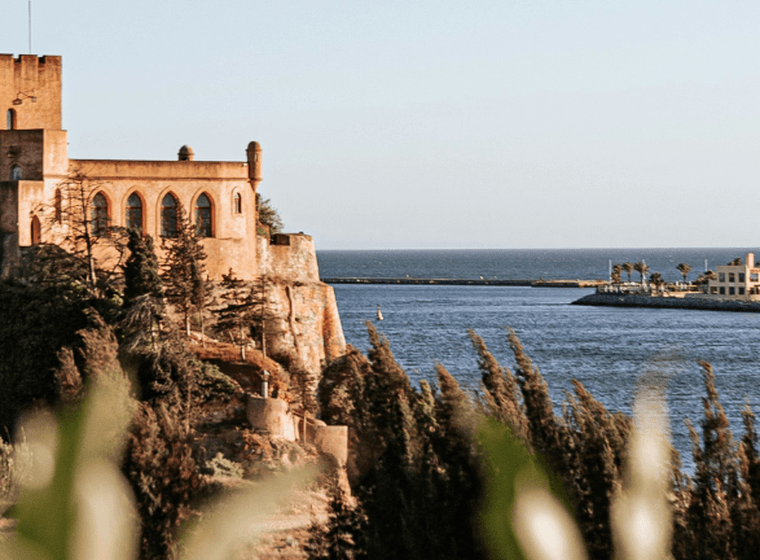 Um castelo histórico numa falésia, com vista para um mar calmo, um porto distante e edifícios sob um céu azul límpido.