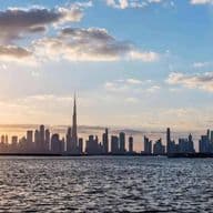Dubai skyline at sunset with tall buildings silhouetted against a colorful sky, reflected in calm water in the foreground.