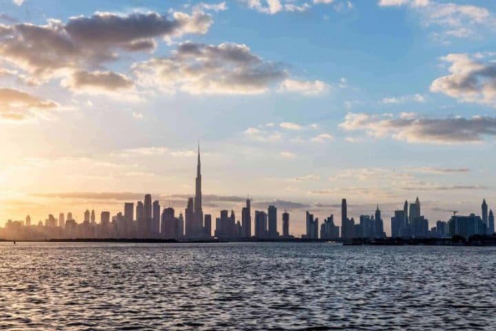 Dubai skyline at sunset with tall buildings silhouetted against a colorful sky, reflected in calm water in the foreground.