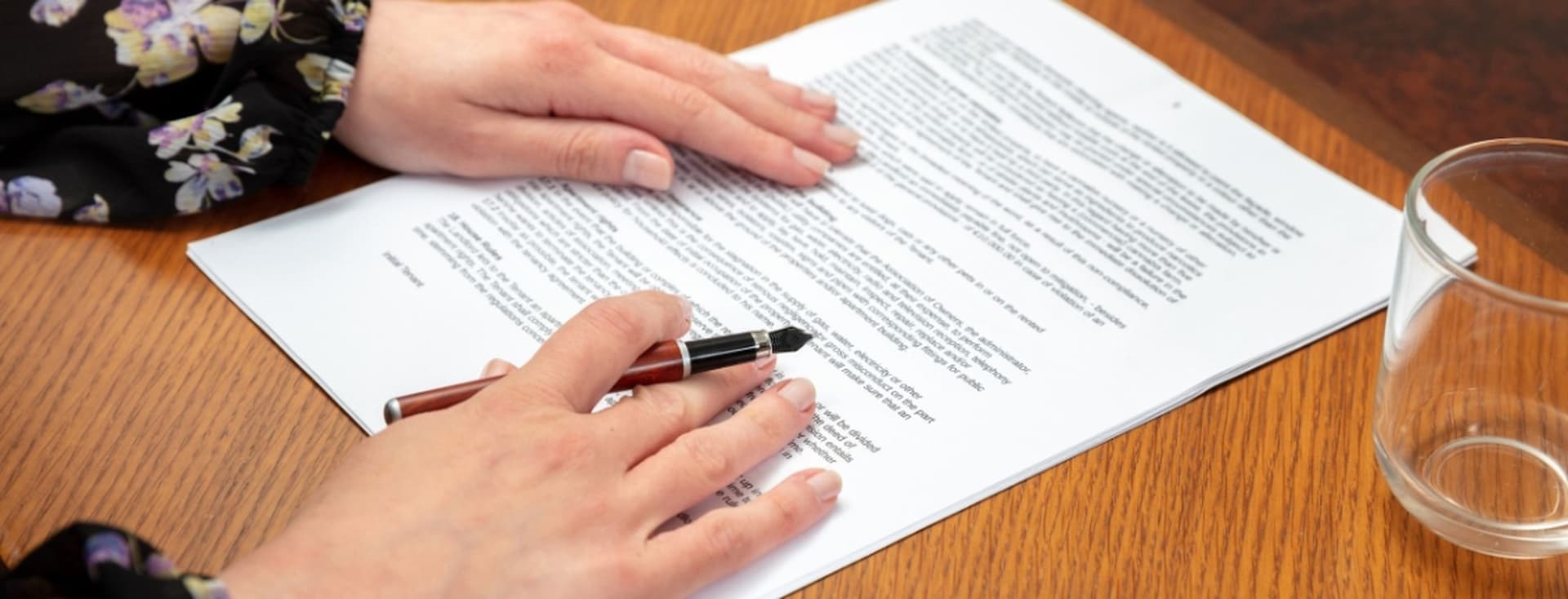 Hände, die einen Stift halten, ruhen auf einem MOU-Dokument auf einem Holztisch, daneben steht ein klares Glas.
