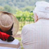 Elderly couple sitting on a bench, wearing hats, facing a scenic view with greenery and trees in the background