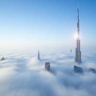 Skyscrapers, including Burj Khalifa, rise above a dense layer of clouds under a clear blue sky.