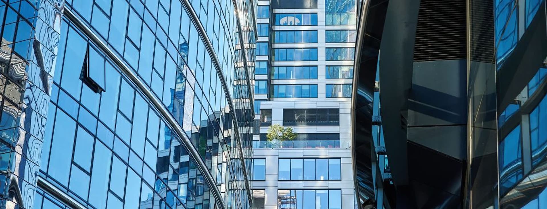 Modern glass buildings reflecting the sky, with a small tree on a balcony, creating a dynamic urban scene.