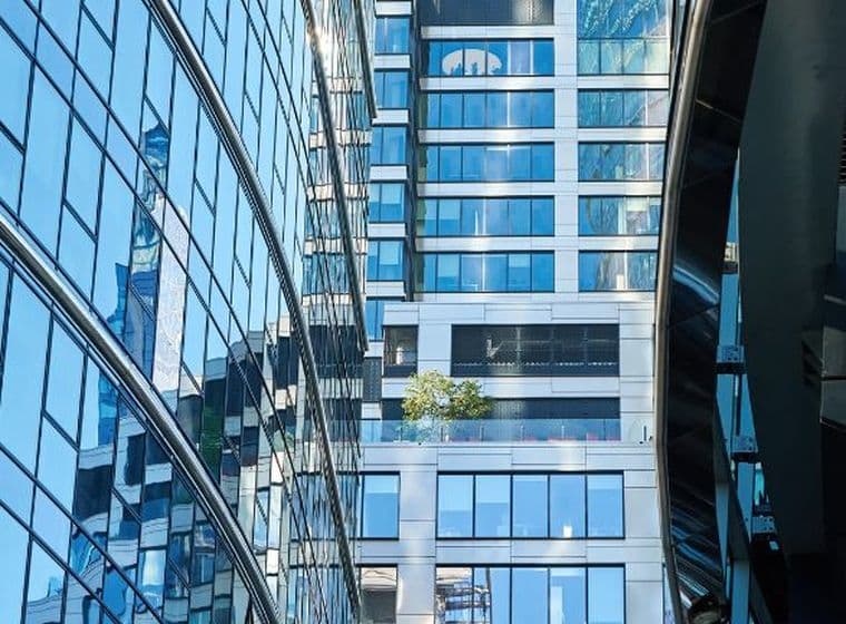 Modern glass buildings reflecting the sky, with a small tree on a balcony, creating a dynamic urban scene.