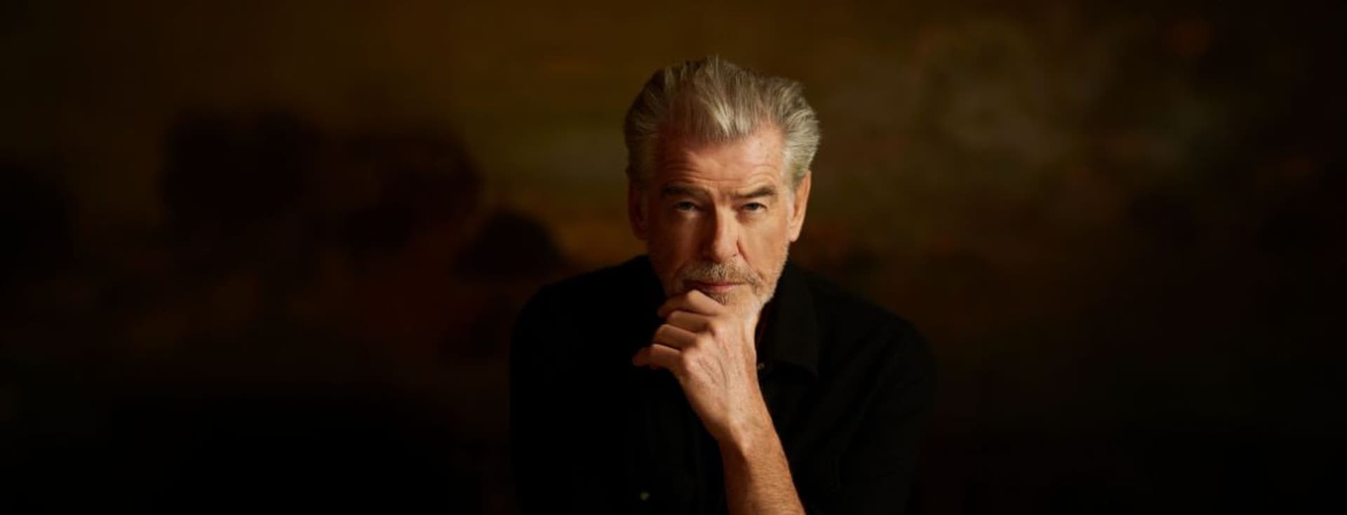 Mann mit grauen Haaren und Bart, der ein schwarzes Hemd trägt und in nachdenklicher Haltung vor einem braunen, unscharfen Hintergrund sitzt.