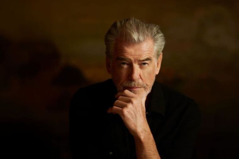 Man with grey hair and beard, wearing a black shirt and sitting in a thoughtful pose in front of a brown, blurred background.