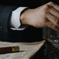 A person in a suit holding a cigar over an ashtray, with an open book and a cigar case nearby.