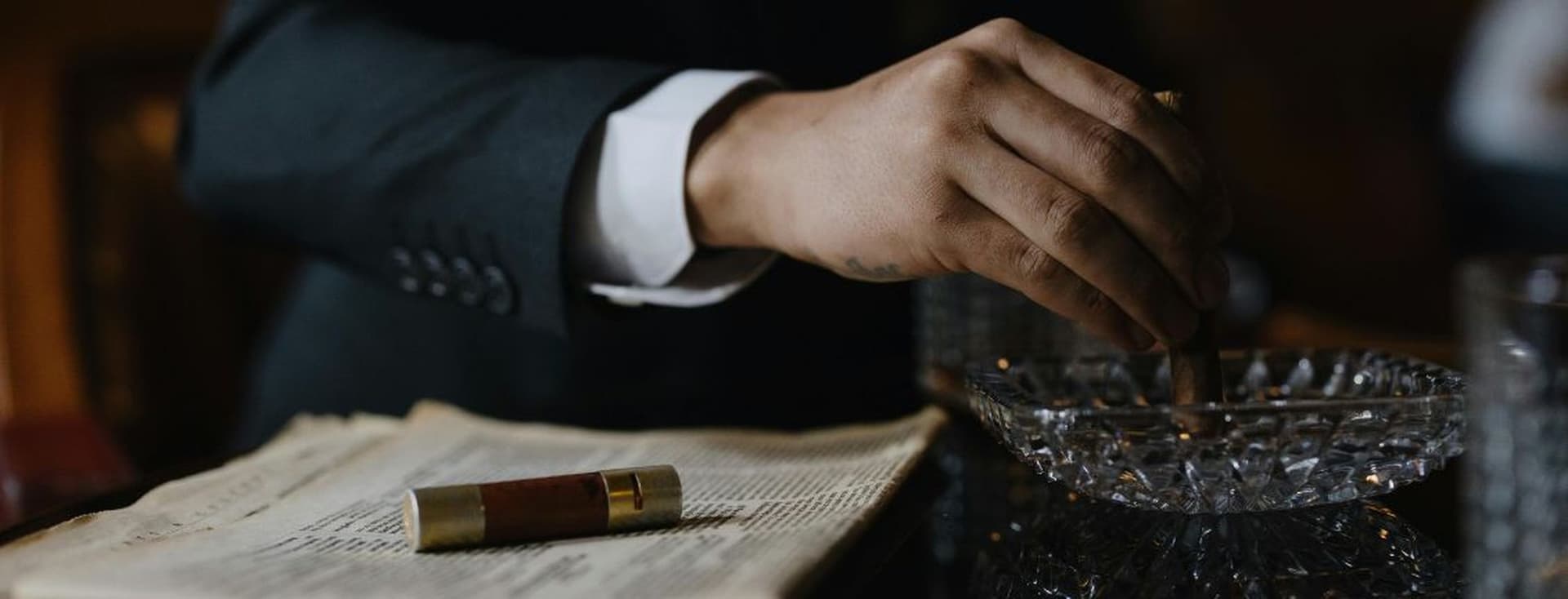 A person in a suit holding a cigar over an ashtray, with an open book and a cigar case nearby.