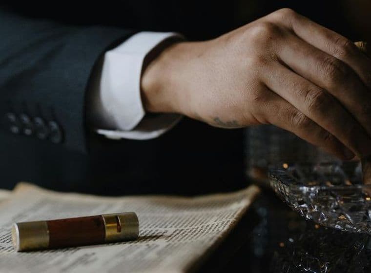 A person in a suit holding a cigar over an ashtray, with an open book and a cigar case nearby.