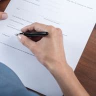 A person signing a document on a wooden desk with a black pen, next to a leather wallet and a set of keys.