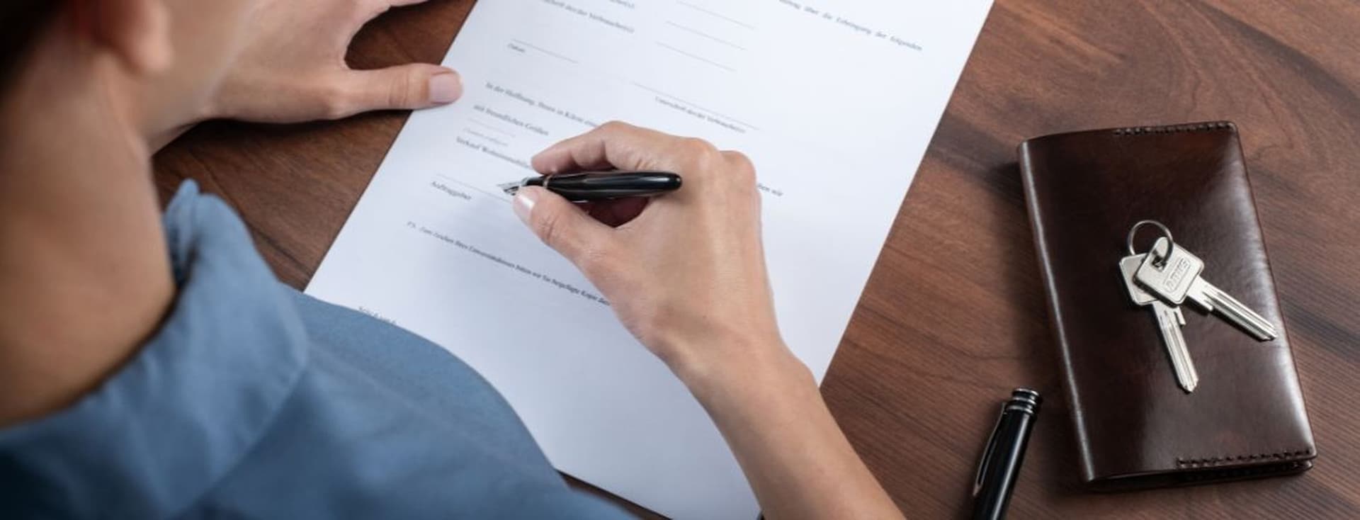 A person signing a document on a wooden desk with a black pen, next to a leather wallet and a set of keys.