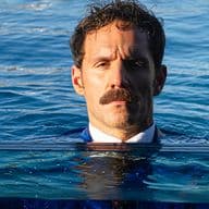 Άντρας με μουστάκι, φορώντας κοστούμι, στέκεται μέσα στο νερό μέχρι το ύψος του στήθους, με φόντο μια ήρεμη γαλάζια θάλασσα.