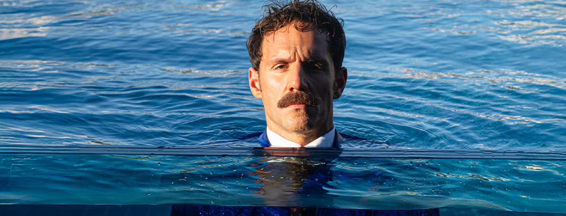Άντρας με μουστάκι, φορώντας κοστούμι, στέκεται μέσα στο νερό μέχρι το ύψος του στήθους, με φόντο μια ήρεμη γαλάζια θάλασσα.
