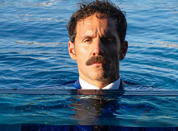 Άντρας με μουστάκι, φορώντας κοστούμι, στέκεται μέσα στο νερό μέχρι το ύψος του στήθους, με φόντο μια ήρεμη γαλάζια θάλασσα.