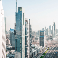 Skyline of DIFC with tall skyscrapers, glass facades, and a wide highway. The scene is bright and clear, showing urban architecture.