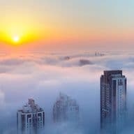 Stadtbild mit Wolkenkratzern, die bei Sonnenaufgang aus einem Wolkenmeer auftauchen, mit heller Sonne und orangefarbenem Himmel.