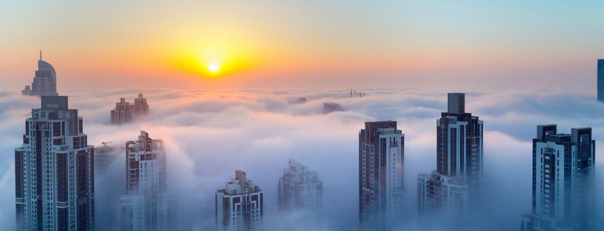 Stadtbild mit Wolkenkratzern, die bei Sonnenaufgang aus einem Wolkenmeer auftauchen, mit heller Sonne und orangefarbenem Himmel.