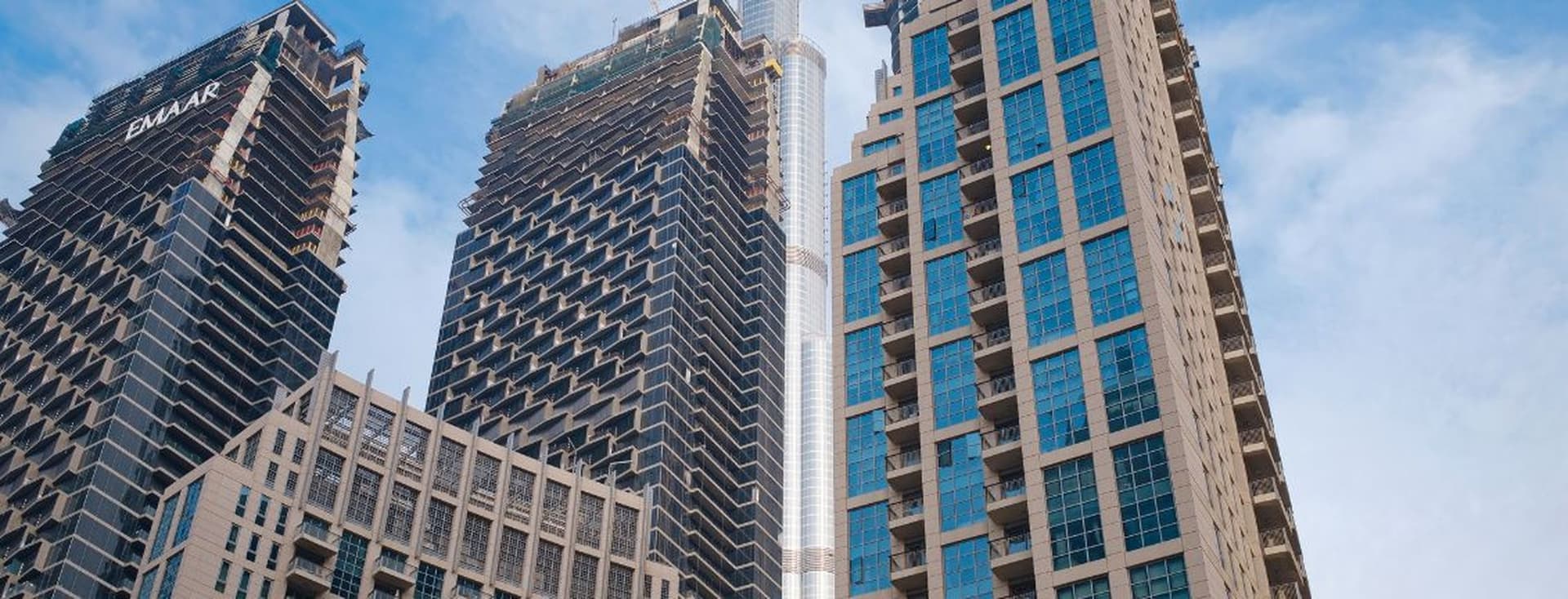 View of modern skyscrapers in Dubai, including the Burj Khalifa, against a cloudy blue sky.