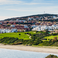 Costa de l'Alcaidesa - vista al mar, la seva platja i la urbanització. Platja en primer terme, camps verds i la urbanització al fons, de cases blanques.