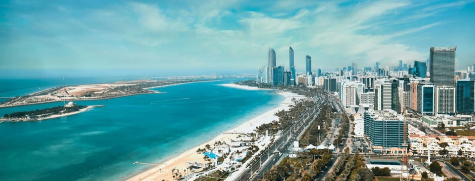 Aerial view of Abu Dhabi with high-rise buildings, a long sandy beach, and blue water under a clear sky.