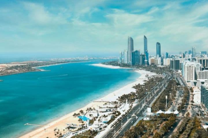 Aerial view of Abu Dhabi with high-rise buildings, a long sandy beach, and blue water under a clear sky.