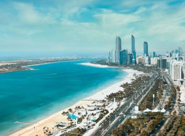 Aerial view of Abu Dhabi with high-rise buildings, a long sandy beach, and blue water under a clear sky.