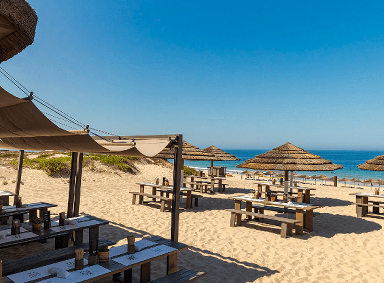 Ein Strandrestaurant mit Holztischen und Strohschirmen am sandigen Ufer, mit Blick auf den blauen Ozean unter einem klaren Himmel.